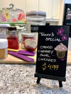 Image of a black mini easel and chalkboard with the words today's special and choco cherry swirl cupcakes written on it and drawing of flowers and cupcake using chalk ink liquid chalk markers standing on a kitchen countertop with cutting board and baking accessories in background