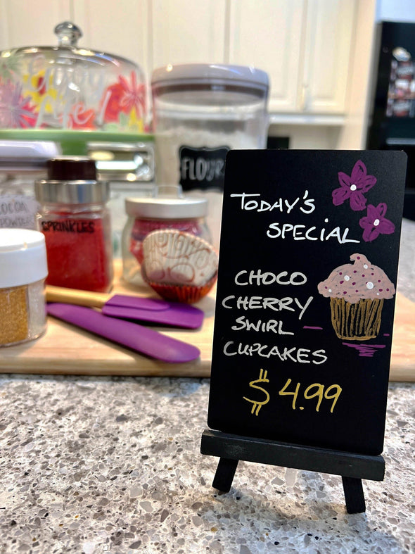 Image of a black mini easel and chalkboard with the words today's special and choco cherry swirl cupcakes written on it and drawing of flowers and cupcake using chalk ink liquid chalk markers standing on a kitchen countertop with cutting board and baking accessories in background
