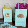 Image of turquoise gift bags with colorful tissue paper and metallic adhesive labels on front of the bags and names written on the labels using chalk ink liquid chalk markers