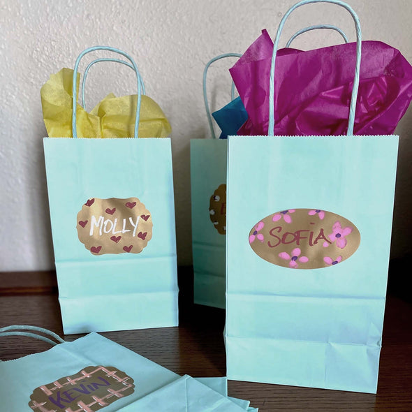 Image of turquoise gift bags with colorful tissue paper and metallic adhesive labels on front of the bags and names written on the labels using chalk ink liquid chalk markers