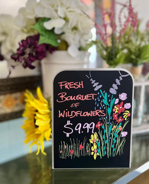 Image of a countertop chalkboard sign in a flower shop with flowers and the words fresh bouquet of wildflowers written on the chalkboard using colorful chalk ink liquid chalk markers  