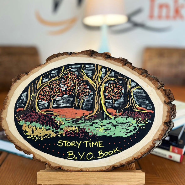 Image of a round wood board decorated with black matte chalkboard paint and a drawn fairytale woods scene and the words story time BYO Book written at the bottom using chalk ink liquid chalk markers
