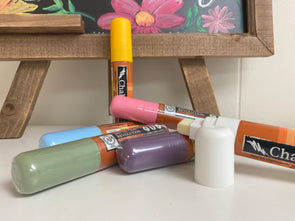 Closeup product image of 15mm bold tip chalk ink liquid chalk markers in spring inspired colors laying on a white table in front of a wood framed chalkboard